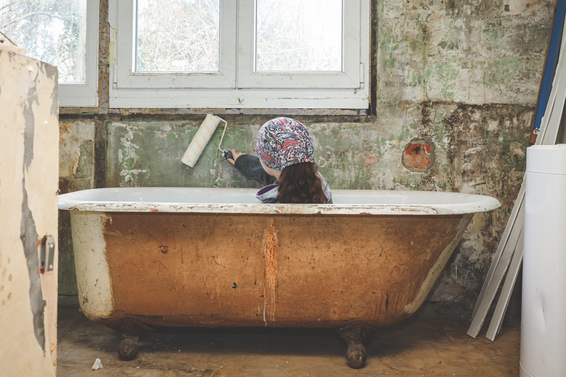 Ein Mädchen sitzt in Arbeitskleidung in der Badewanne und streicht eine Wand